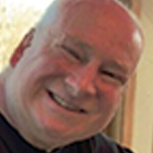 A middle-aged man with a bald head smiles warmly at the camera. He is indoors, and soft light is coming from the background.