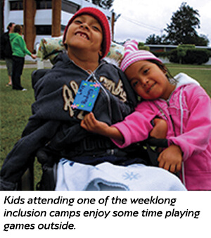 Two children, one in a wheelchair, smile and pose together outside at a camp. The girl hugs the boy, and both wear warm clothes and hats. Other people and buildings are visible in the background.