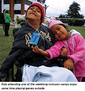 Two smiling children, one in a wheelchair and the other hugging them, enjoy outdoor games at an inclusion camp. Both are dressed warmly, with a green lawn and buildings in the background.