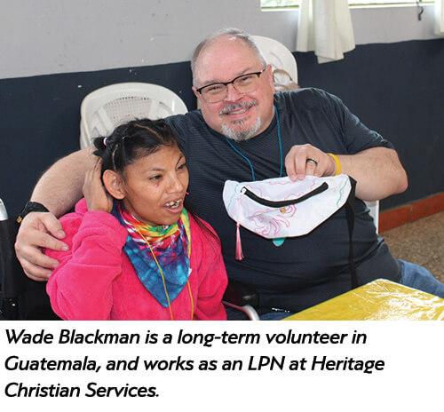 A smiling man with glasses sits beside a young girl in a wheelchair. They are indoors at a table. The text says he is Wade Blackman, a long-term volunteer in Guatemala and an LPN at Heritage Christian Services.