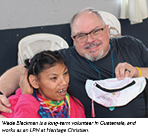 A smiling man with glasses and a beard hugs a young girl with special needs. They are sitting together on white chairs. The man holds a white bag, and both appear happy.