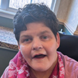 A woman with short, curly dark hair smiles, wearing a pink patterned scarf. She is indoors, seated near a window with sunlight coming through.