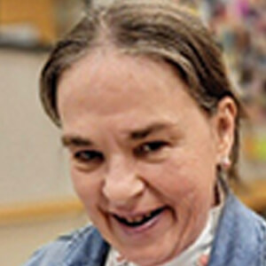 A woman with light skin and graying brown hair smiles at the camera. She is indoors, wearing a blue jacket and a light-colored top. The background is slightly blurred.