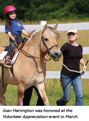 A smiling young girl wearing a helmet rides a light brown horse, led by a woman in a cap along a fenced path. The scene takes place outdoors on a sunny day, with green grass and trees in the background.