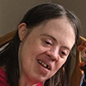 A person with long brown hair and a pink shirt sits and smiles, looking down. The photo is taken indoors with warm lighting and a wooden chair is visible in the background.