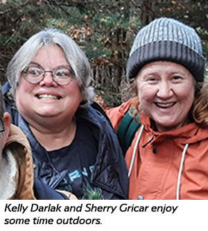 Two smiling women, one wearing glasses and a dark jacket, the other in a rust-colored jacket and knit hat, stand close together outdoors with trees in the background.