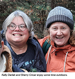 Two women dressed warmly in jackets and hats smile while standing close together outdoors, surrounded by trees.