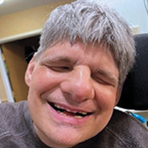 A person with short gray hair is smiling widely while sitting indoors, wearing a brown shirt and seated in a wheelchair.