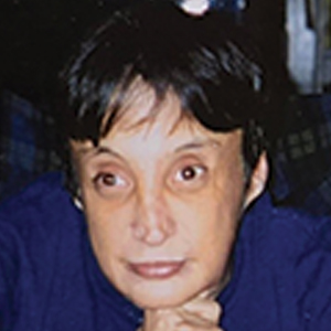 A person with short dark hair and a thoughtful expression rests their chin on their hand, wearing a dark blue top, against a dark, blurred background.