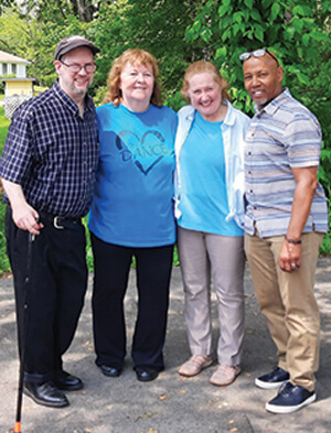 Four adults stand closely together outdoors, smiling at the camera. Trees and greenery are in the background. One man holds a cane, and the group appears friendly and happy.