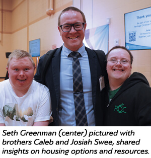 Three smiling people pose indoors. The man in the center wears glasses, a suit, and tie. On each side, two men in casual clothing stand close. Informational signs and a “Thank you” message are visible in the background.