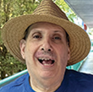 A man wearing a wide-brimmed straw hat and blue shirt smiles outdoors, with green trees and a walkway in the background.