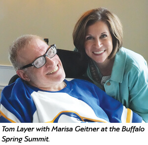 A smiling man in a blue and white sports jersey sits in a wheelchair next to a woman with short brown hair, who is also smiling. They are at the Buffalo Spring Summit.