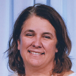 A woman with shoulder-length brown hair is smiling in front of a light blue curtain background.