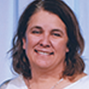 A woman with medium-length brown hair, wearing a white top, smiles at the camera against a softly blurred, light-colored background.