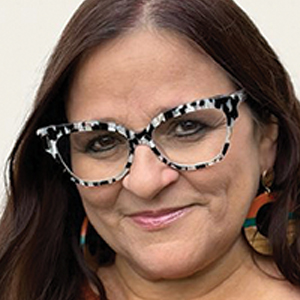 A woman with long brown hair wears large, black-and-white patterned glasses and colorful round earrings. She is smiling slightly and standing in front of a light-colored background.