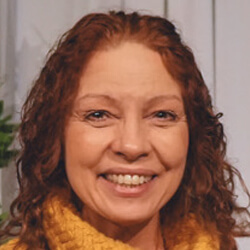 A woman with curly red hair and a warm smile wearing a yellow sweater, standing indoors in front of a light-colored wall.