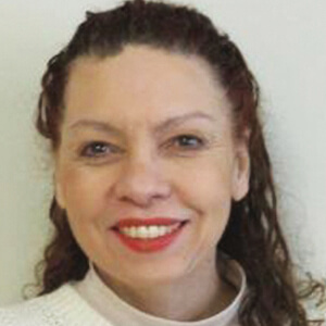 A woman with wavy brown hair, wearing a white top, smiles at the camera against a plain light background.