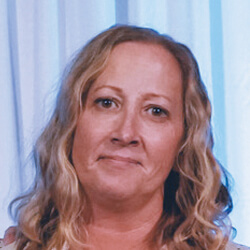 A woman with wavy blonde hair and light skin stands in front of white curtains, looking at the camera with a gentle expression.