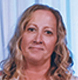 A woman with wavy blonde hair and a light complexion is standing in front of a blurred background with light curtains, looking at the camera with a neutral expression.
