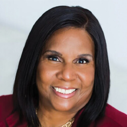 A woman with straight, shoulder-length dark hair smiles warmly at the camera. She is wearing a burgundy blazer and a gold necklace, with a light-colored background behind her.