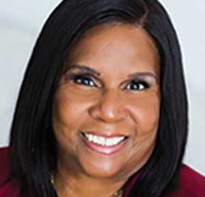 A woman with straight, shoulder-length dark hair smiles warmly at the camera. She is wearing a dark-colored top and is pictured against a plain, light background.