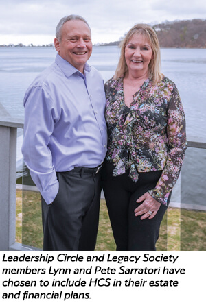 A smiling man and woman stand together by a lakeside railing, dressed in business-casual clothing. The caption notes they are Lynn and Pete Sarratori, who have included HCS in their estate and financial plans.
