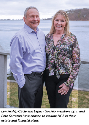 A smiling man and woman stand side by side on a deck overlooking a lake. The man wears a light purple shirt and dark pants; the woman wears a floral blouse and black pants. Trees and water are visible in the background.