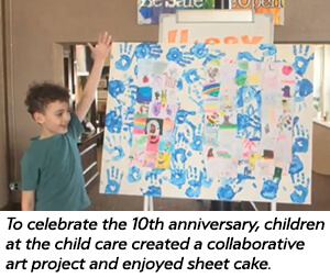 A young child stands beside a large art board decorated with blue handprints and drawings, celebrating the 10th anniversary of a child care center. A caption explains the collaborative art project and a celebration with sheet cake.