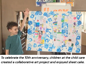 A young child stands beside a large poster covered with colorful drawings and blue handprints. A caption notes the artwork celebrates the child cares 10th anniversary.