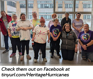 A group of people pose and smile together indoors in front of large windows. Some wear colorful tie-dye shirts, others in purple shirts. Text below reads: Check them out on Facebook at tinyurl.com/HeritageHurricanes.