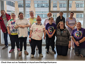 A group of ten people, some wearing colorful tie-dye shirts and others in solid tops, stand smiling indoors in front of large windows with buildings visible outside.