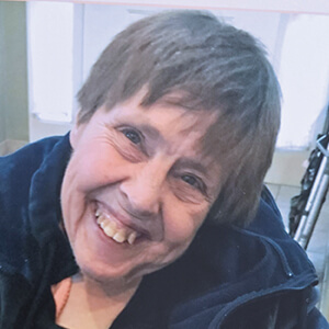 A woman with short brown hair smiles warmly at the camera, wearing a dark jacket. The background is indoors with bright light coming from windows behind her.