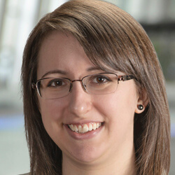 A woman with straight brown hair, wearing glasses and small black earrings, is smiling. The background is softly blurred, suggesting an indoor setting with light coming from the left.