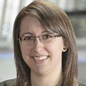 A woman with straight, shoulder-length brown hair and glasses smiles at the camera. She is wearing a grey top, and the background is softly blurred, suggesting an indoor setting.