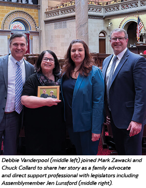 Four people stand and smile in a formal government chamber. One woman holds a framed photo. They are dressed in business attire, with ornate arches and American flags visible in the background.