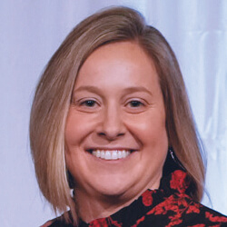 A woman with straight, shoulder-length blonde hair smiles at the camera. She is wearing a black top with a red floral pattern and is posed in front of a light-colored background.
