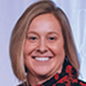 A woman with straight, shoulder-length blonde hair smiles at the camera. She is wearing a black top with a red floral pattern. The background is light and slightly blurred.