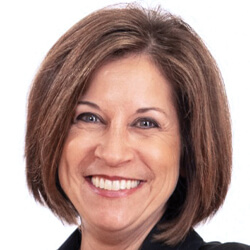 A woman with short brown hair smiles at the camera. She is wearing a black top and is photographed against a plain white background.