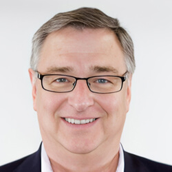 A middle-aged man with short gray hair and glasses is smiling at the camera. He is wearing a dark blazer over a light-colored shirt and is posed against a plain white background.
