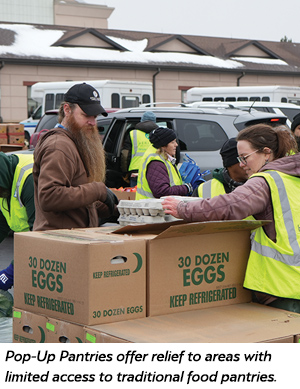 Volunteers in yellow vests distribute cartons of eggs from large boxes at an outdoor pop-up pantry event, with vans and buildings visible in the background.