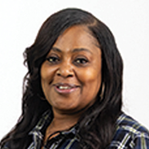 A woman with long, wavy dark hair smiles at the camera. She is wearing a plaid shirt and large hoop earrings, standing in front of a plain white background.