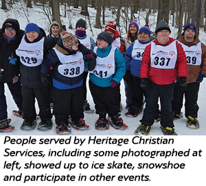 A group of people standing outdoors in the snow, wearing winter clothes, numbered race bibs, and snowshoes, posing together and smiling for the camera. Trees are visible in the background.