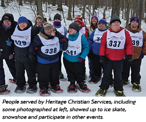 A group of people in winter clothing and numbered vests stand together outdoors in the snow, smiling and wearing snowshoes, with trees in the background.