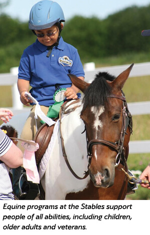 A young child wearing a helmet and glasses smiles while sitting on a brown and white horse, holding a ribbon. Adults assist nearby. Text at bottom highlights equine programs supporting people of all abilities.