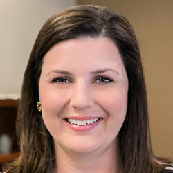 A woman with straight brown hair smiles at the camera. She is indoors, and the background is softly blurred, suggesting an office or home setting.