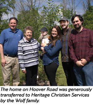 Six people, including a baby, stand outdoors smiling for a group photo. An older woman and man are on the left, with younger adults and a baby in the center and right. Trees and green foliage are in the background.