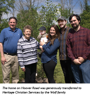 Six people of varying ages stand together outside on green grass with trees in the background, smiling at the camera. A woman holds a baby. A caption notes a home transfer to Heritage Christian Services by the Wolf family.
