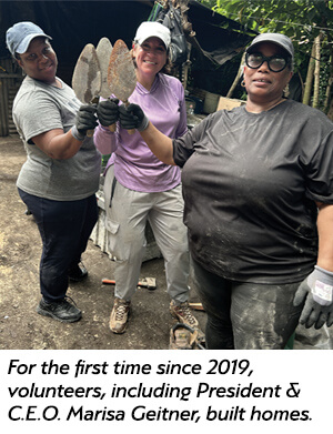 Three women volunteers, smiling and holding masonry trowels, pose together at a construction site. They wear work gloves and casual clothes, with building materials visible in the background.