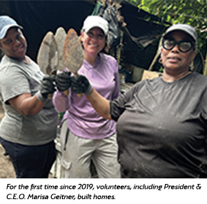 Three smiling women, wearing work clothes and gloves, stand outdoors and hold up trowels together. In the background are trees and construction materials. A caption notes they are volunteers building homes.
